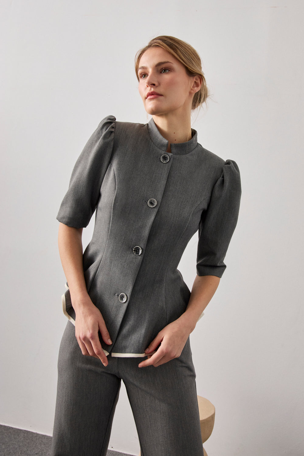 Chaqueta entallada en color gris con vuelo y pinzas en pecho. Tiene hombros rizados, manga corta, cuello mao y detalle a contraste en bajo.